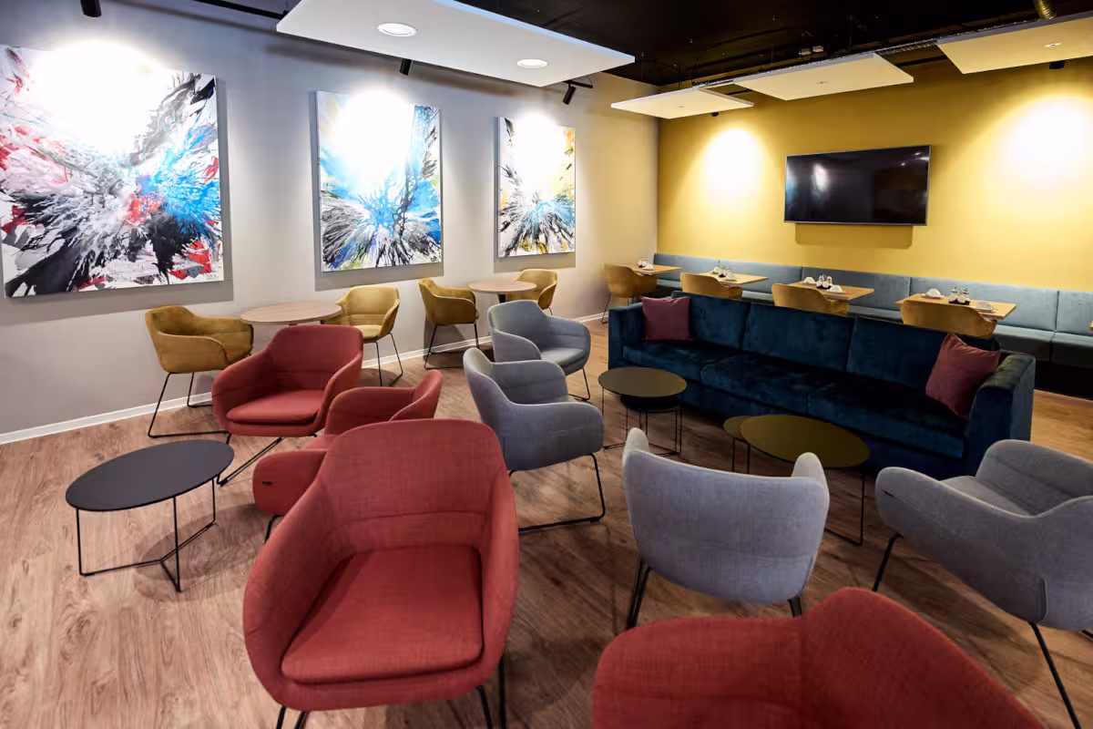 Eine moderne Lounge mit roten und grauen Sesseln, runden schwarzen Tischen, Wandkunst, einem dunkelblauen Sofa, einem Flachbildfernseher und Holzfußboden. Gelbe und weiße Wände sind sichtbar, mit Deckenpaneelen und eingelassener Beleuchtung.