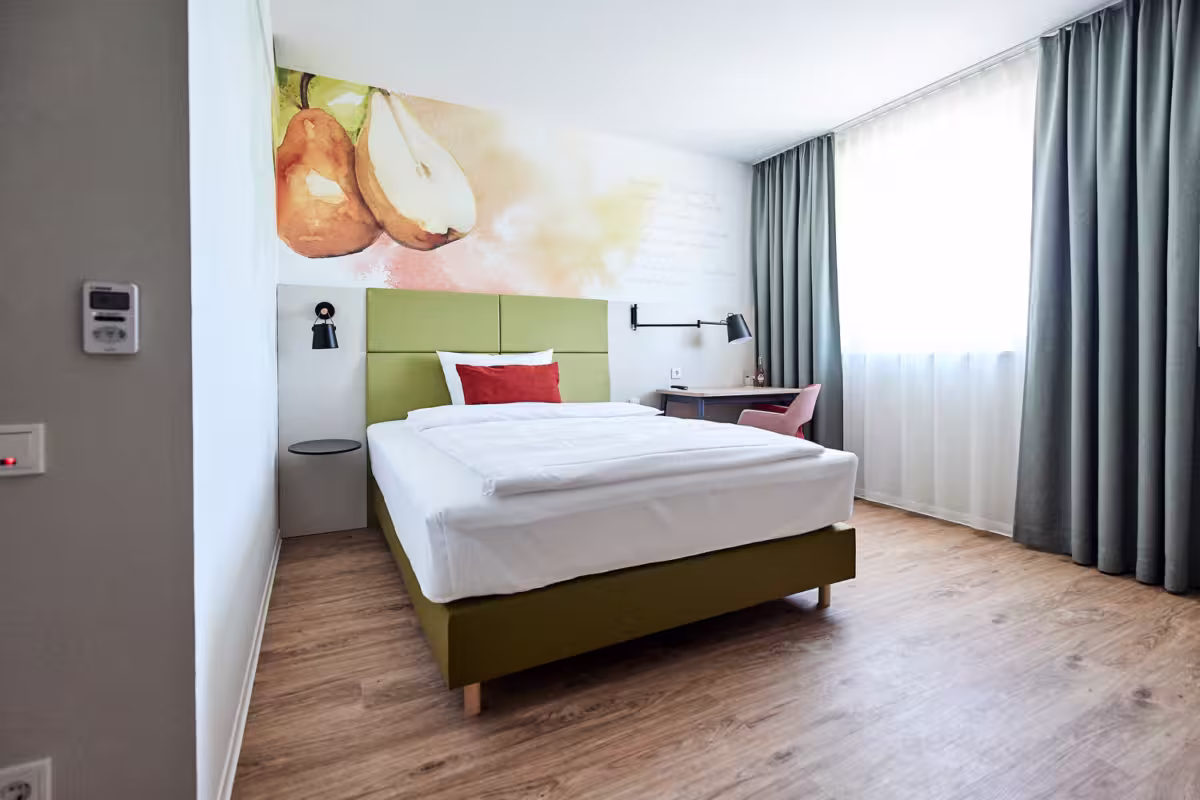 Ein modernes Hotelzimmer mit einem Einzelbett, grünem Kopfteil, weißem Bettzeug und einem roten Akzent-Kissen. Ein Schreibtisch und ein Stuhl stehen am Fenster. An der Wand hängt ein großes Wandbild mit Birnen und Äpfeln. Das Zimmer hat einen Holzfußboden und graue Vorhänge.