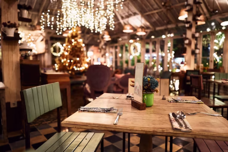 Ein warm beleuchtetes Restaurant mit Holztischen für zwei Personen, festlichen Lichterketten an der Decke und einem geschmückten Weihnachtsbaum im Hintergrund, der eine gemütliche und einladende Atmosphäre schafft. Business-Fotografie Mannheim, Heidelberg und Ludwigshafen