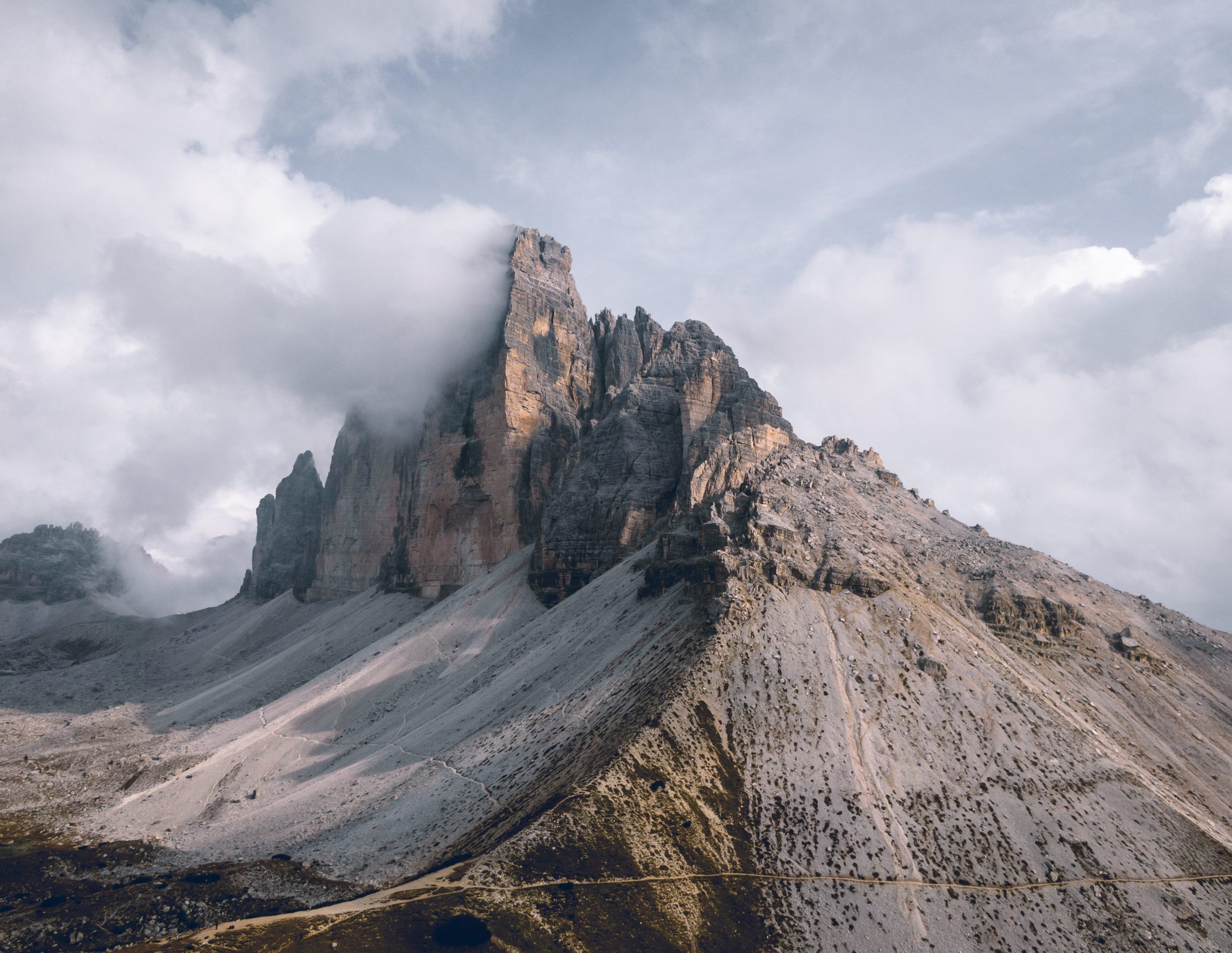 Ein felsiger Berggipfel erhebt sich steil und ist in Gipfelnähe teilweise von Wolken bedeckt. Die Hänge sind steil und karg, mit verstreuten Felsen und einem wolkenverhangenen Himmel über uns. Es gibt kaum sichtbare Vegetation.