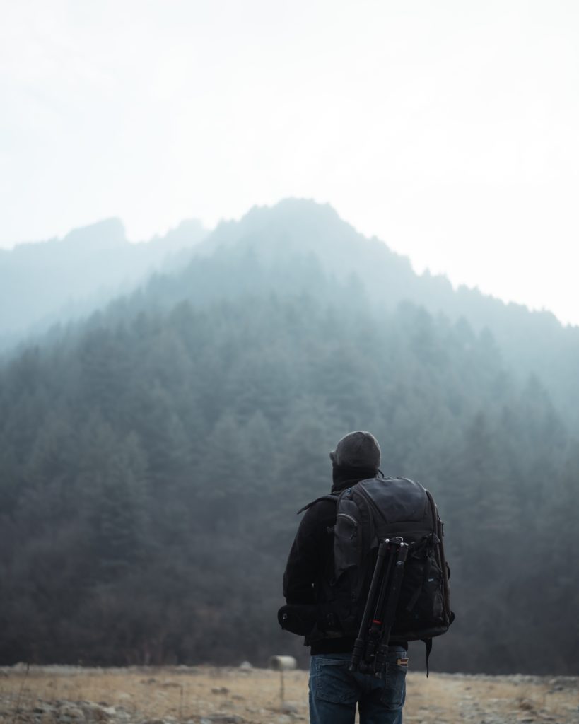 Eine Person, die eine dunkle Jacke und eine Mütze trägt, steht im Freien und blickt auf einen nebligen, baumbedeckten Berg. Die Person hat einen großen Rucksack mit einem Stativ dabei und steht auf trockenem Gras.