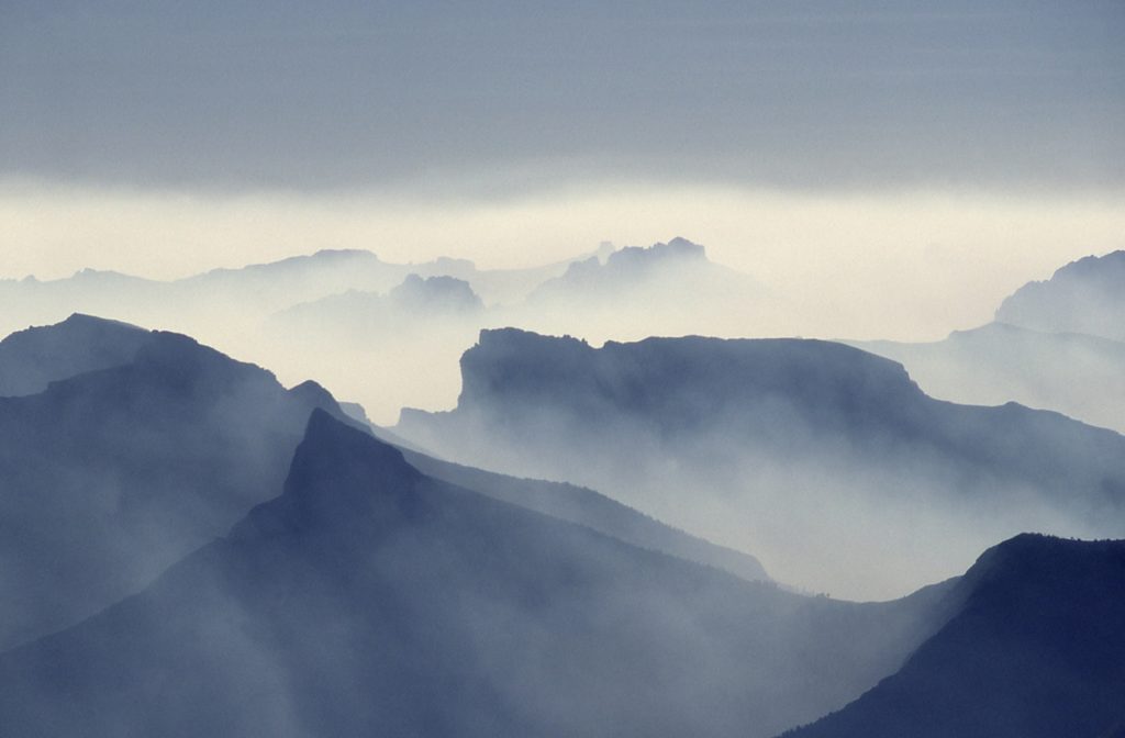 Schichten von Gebirgskämmen verschwinden in der Ferne, teilweise verdeckt von Dunst und Nebel unter einem bewölkten Himmel, wodurch eine weiche, blaugraue Atmosphäre entsteht.